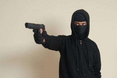 Siyah kapüşonlu ve maskeli gizemli bir adam tabanca tutuyor, silahla ateş ediyor. Gri arkaplanda izole edilmiş resim