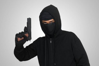 Siyah kapüşonlu ve maskeli gizemli bir adam tabanca tutuyor, silahla ateş ediyor. Gri arkaplanda izole edilmiş resim
