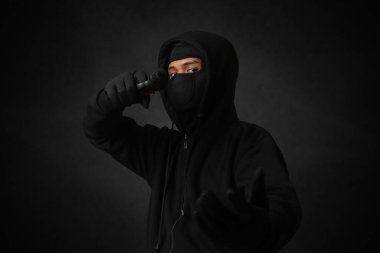 Siyah kapüşonlu ve maskeli gizemli bir adam tabanca tutuyor, silahla ateş ediyor. Karanlık ortam arkaplanındaki izole görüntü