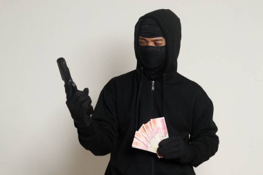 Siyah kapüşonlu ve maskeli gizemli bir adamın portresi. Elinde 100 bin rupiah banknot tutarken silahla ateş ediyor. Para sızdırıyor. Gri arkaplanda izole edilmiş resim