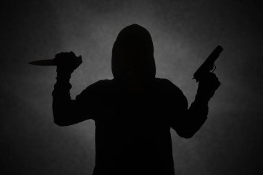 Siyah kapüşonlu, tabanca tutan gizemli bir adam. Elinde bıçak tutarken silahla ateş ediyor. Siluet ve karanlık konsept görüntüsü