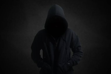 Siyah kapüşonlu gizemli adam karanlık arka planda duruyor. Hacker, suç ve siber güvenlik kavramı.
