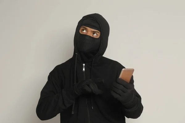 神秘男子身穿黑色帽衫 戴着面具在手机上做黑客活动 黑客手持智能手机的肖像 灰色背景上的孤立图像 — 图库照片