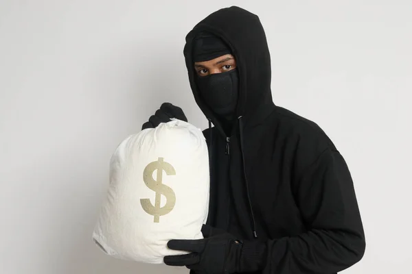 黒いパーカーを着た謎の泥棒男とお金でいっぱいの袋を運ぶマスク 灰色の背景に孤立した画像 — ストック写真