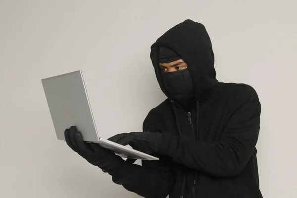 stock image Portrait of mysterious man wearing black hoodie and mask doing hacking activity on laptop, hacker holding a personal computer. Cyber security concept. Isolated image on gray background