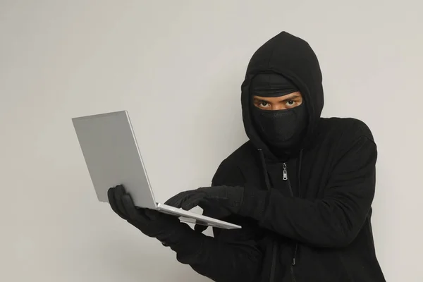 Portrait of mysterious man wearing black hoodie and mask doing hacking activity on laptop, hacker holding a personal computer. Cyber security concept. Isolated image on gray background