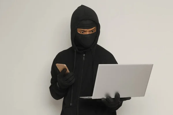 黒いパーカーとマスクを身に着けている謎の男の肖像画は ノートパソコンでハッキング活動を行い ハッカーは携帯電話を保持しています サイバーセキュリティの概念 灰色の背景に孤立した画像 — ストック写真