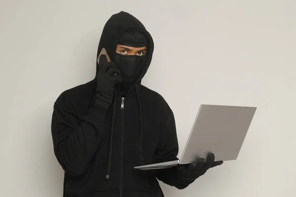 stock image Portrait of mysterious man wearing black hoodie and mask doing hacking activity on laptop, hacker holding a mobile phone. Cyber security concept. Isolated image on gray background