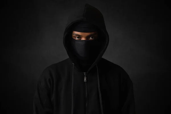 Mysterious man wearing black hoodie and mask standing against dark background, looking at camera. Dramatic low light portrait