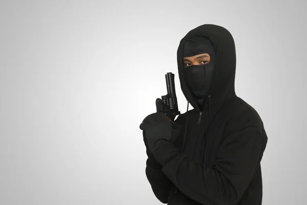 黒いパーカーを着た謎の男とピストルを持ってマスク銃で撃つ 灰色の背景に孤立した画像 — ストック写真