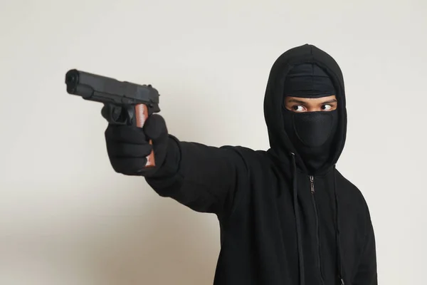 黒いパーカーを着た謎の男とピストルを持ってマスク銃で撃つ 灰色の背景に孤立した画像 — ストック写真