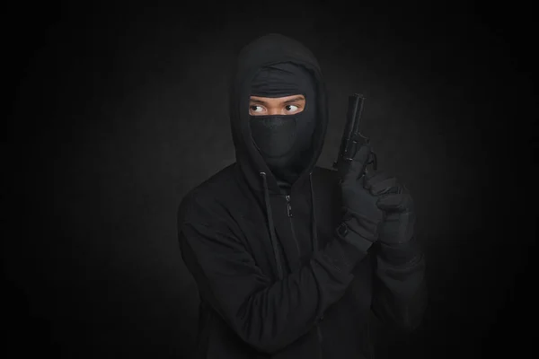 黒いパーカーを着た謎の男とピストルを持ってマスク銃で撃つ 暗い周囲の背景に隔離された画像 — ストック写真