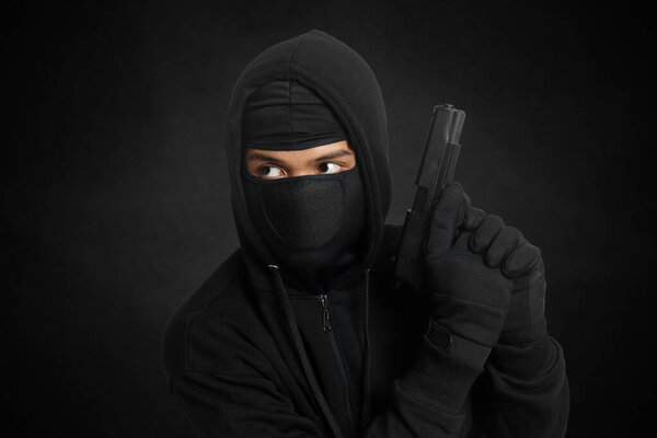 Mysterious man wearing black hoodie and mask holding a pistol, shooting with a gun. Isolated image on dark ambient background