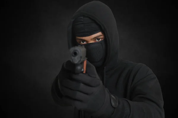 黒いパーカーを着た謎の男とピストルを持ってマスク銃で撃つ 暗い周囲の背景に隔離された画像 — ストック写真