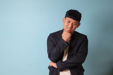 Asyalı genç iş adamının portresi. Sıradan bir takım elbiseyle düşünürken gülümsüyor. Mavi arkaplanda izole edilmiş resim
