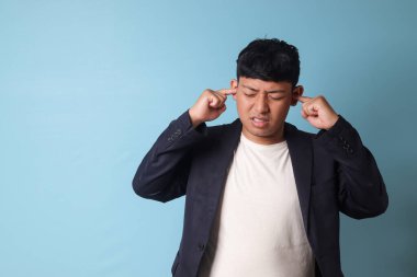 Sıradan giyinmiş Asyalı genç iş adamının portresi iki eliyle kulaklarını kapatırken ve gürültü içinde ezilirken. Mavi arkaplanda izole edilmiş resim
