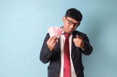 Binlerce rupia tutarken kameraya bakıp işaret eden sıradan takım elbiseli Asyalı genç iş adamının portresi. Mavi arkaplanda izole edilmiş resim