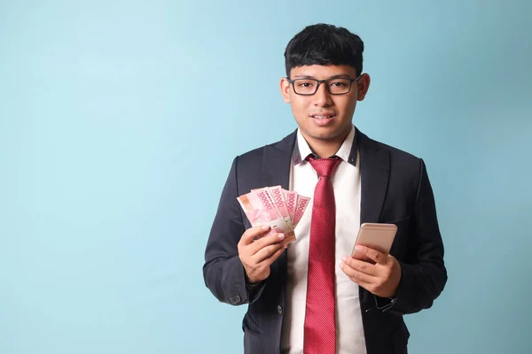 stock image Portrait of young Asian business man in casual suit holding phone and thousand rupiahs looking at camera smiling. Isolated image on blue background