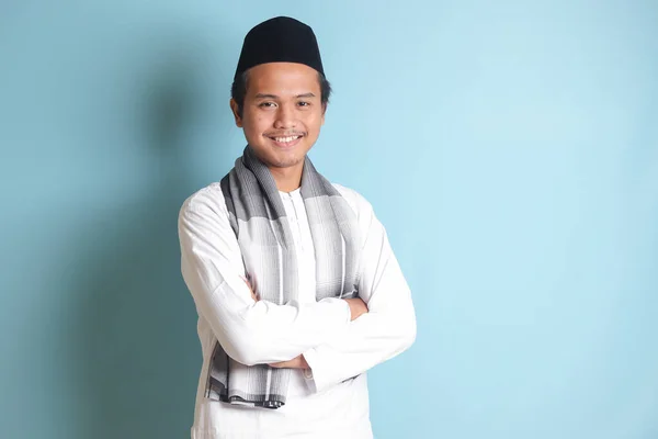 Portrait of religious Asian man in koko shirt or white muslim shirt and black cap, standing with crossed arms and looking at camera. Isolated image on blue background