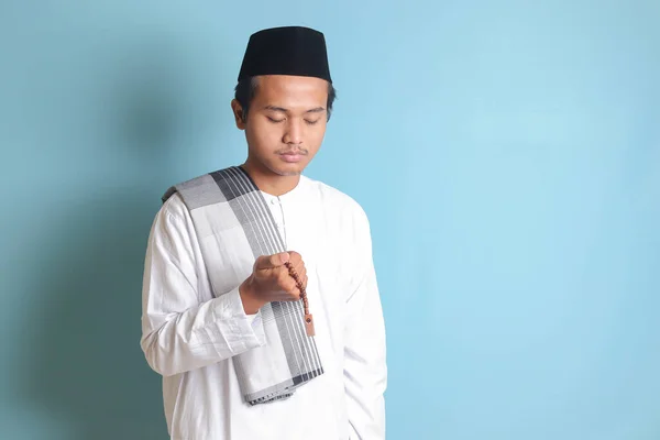 亚洲穆斯林男子的画像 身穿白色科科衬衫 头戴便帽 双手高举 诚挚地祈祷 蓝色背景上的孤立图像 — 图库照片