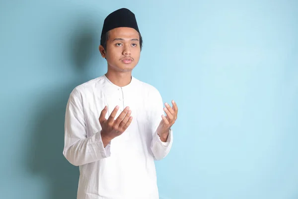 亚洲穆斯林男子的画像 身穿白色科科衬衫 头戴便帽 双手高举 诚挚地祈祷 蓝色背景上的孤立图像 — 图库照片