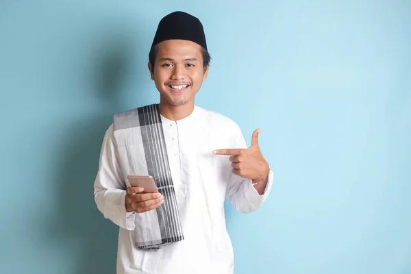 一个年轻的亚洲穆斯林男子拿着手机 面带微笑 手指指向旁边的画像 蓝色背景上的孤立图像 — 图库照片