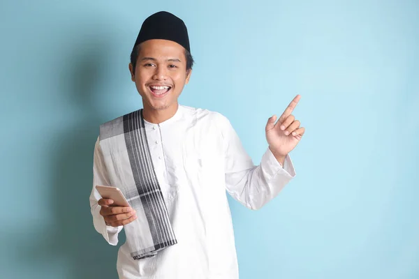 一个年轻的亚洲穆斯林男子拿着手机 面带微笑 手指指向旁边的画像 蓝色背景上的孤立图像 — 图库照片