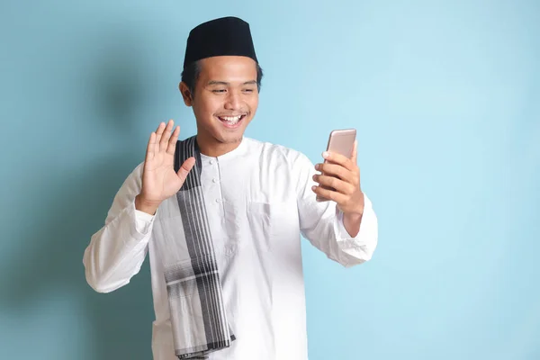 亚洲穆斯林男子的画像 身穿白色科科衬衫 头戴小帽 为自己或自己拍照 在视频通话中打招呼和挥手 蓝色背景上的孤立图像 — 图库照片