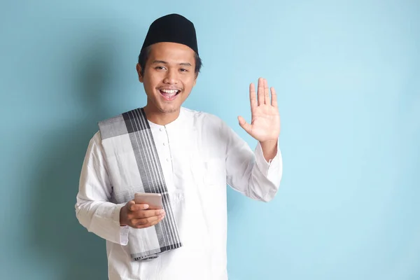年轻的亚洲穆斯林男子面带微笑地拿着手机 挥挥手向摄像机致意的画像 蓝色背景上的孤立图像 — 图库照片