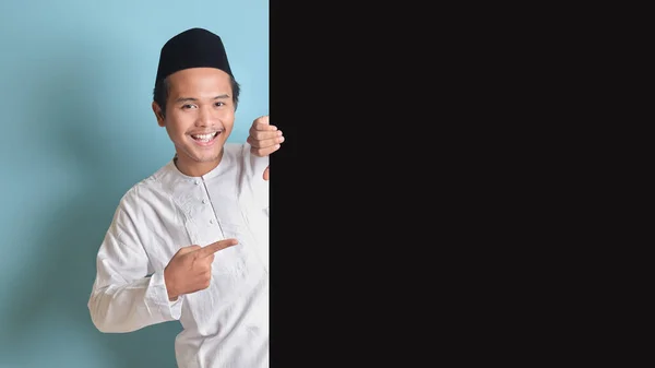 あなたのテキストを宣伝する大きな黒い紙のポスターのコピースペースで製品と指差し指を示す若いアジアのイスラム教徒の男性の肖像画 青い背景に孤立したイメージ — ストック写真