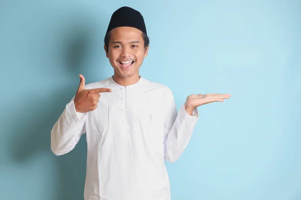 年轻的亚洲穆斯林男子的画像 展示产品 用他的手和手指指向侧面 蓝色背景上的孤立图像 — 图库照片