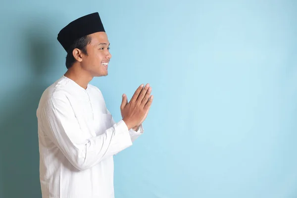 謝罪と歓迎の手のジェスチャーを示すSkullcapと白いココシャツのアジアのイスラム教徒の男の肖像画 青い背景に孤立したイメージ — ストック写真