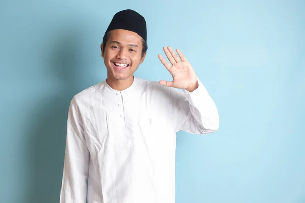 亚洲穆斯林男子的画像 身穿白色可可衬衫 头戴便帽 向人打招呼 挥手拍照 面带微笑地向人打招呼 蓝色背景上的孤立图像 — 图库照片