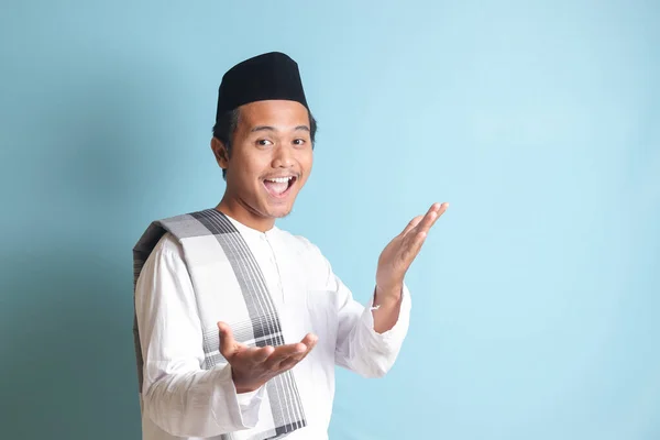 亚洲穆斯林男子的画像 身穿白衣 头戴便帽 手牵着相机 邀请人进来 欢迎大家做手势 蓝色背景上的孤立图像 — 图库照片