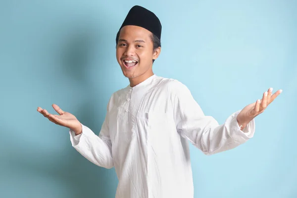 亚洲穆斯林男子的画像 身穿白衣 头戴便帽 手牵着相机 邀请人进来 欢迎大家做手势 蓝色背景上的孤立图像 — 图库照片