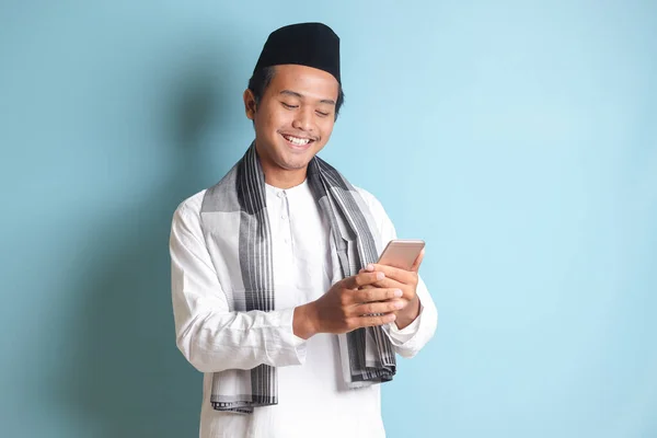 年轻的亚洲穆斯林男子面带微笑地拿着和触摸着手机的画像 蓝色背景上的孤立图像 — 图库照片