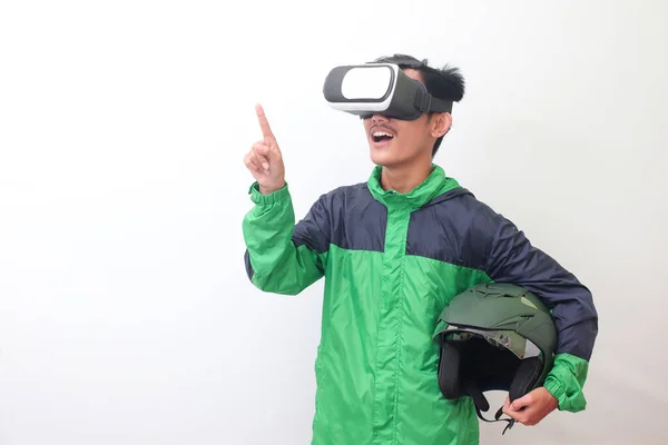 Portrait of Asian online taxi driver wearing green jacket and holding a helmet while using virtual reality or VR and pointing. Isolated image on white background