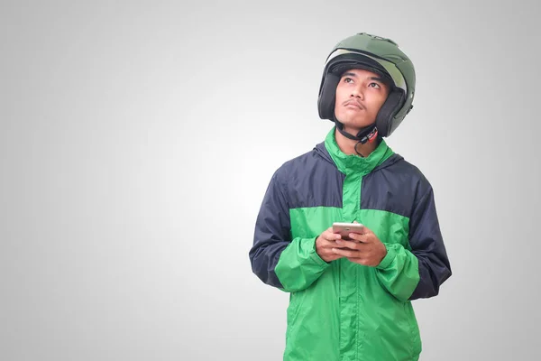 Portrait of confused Asian online taxi driver wearing green jacket and helmet thinking about an idea and looking up. Isolated image on white background