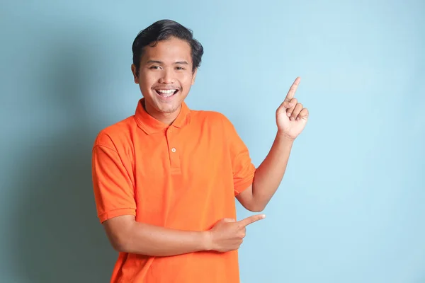 身穿橙色衬衫的兴奋的亚洲男人笑着看着摄像机 用双手和手指指向旁边 蓝色背景上的孤立图像 — 图库照片