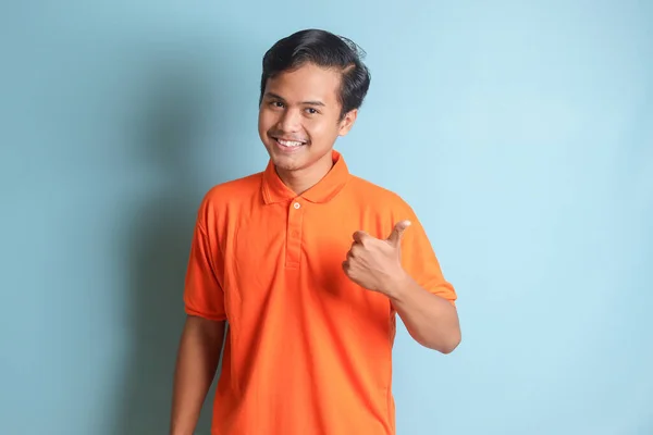 空の空間に親指を向けてオレンジのシャツの魅力的なアジア人男性 広告コンセプト 青い背景に孤立したイメージ — ストック写真