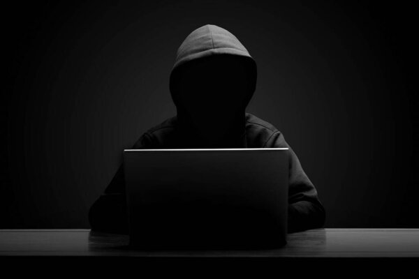 Таинственный человек в черной толстовке с ноутбуком в темной комнате. Концепция хакера и кибербезопасности.
