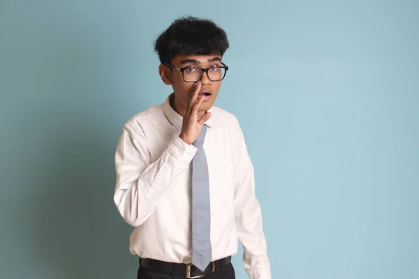 Indonesian senior high school student wearing white shirt uniform with gray tie whispering malicious talk conversation, hand on mouth telling secret rumor. Isolated image on gray backgroundIndonesian senior high school student wearing white shirt uni