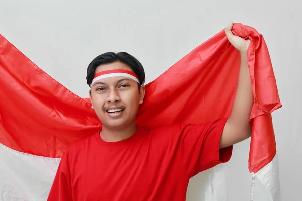 赤と白のリボンを頭にしたTシャツを着た魅力的なアジア人男性の肖像画は 彼の拳で旗を掲げ インドネシアの独立記念日を祝います 灰色の背景に孤立した画像 — ストック写真