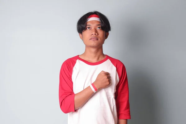 Portrait of attractive Asian man in t-shirt with red and white ribbon on head, placing his hand on chest, giving respect and salute hand gesture. Isolated image on gray background