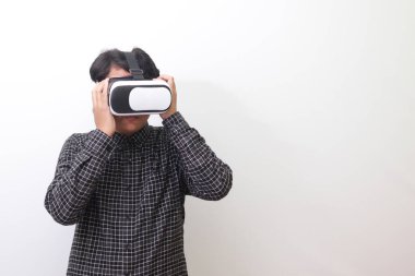 Siyah ekose gömlekli Asyalı bir adamın Sanal Gerçeklik (VR) gözlüklerini takıp yukarı bakarken portresi. Beyaz arkaplanda izole edilmiş resim
