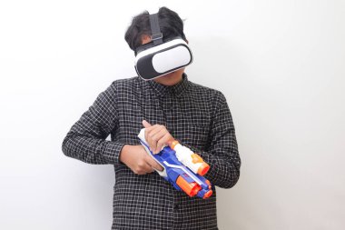 Siyah ekose gömlekli Asyalı bir adamın Sanal Gerçeklik (VR) gözlüklerini kullanarak bir silah silahı konsolu tutarken portresi. Beyaz arkaplanda izole edilmiş resim