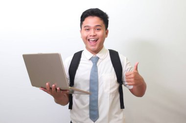 Endonezyalı lise öğrencisi beyaz gömlekli, gri kravatlı, dizüstü bilgisayarlı ve el hareketlerini gösteren gri arka planda duran.