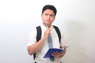 Endonezyalı lise öğrencisi beyaz gömlekli, gri kravatlı kalemle not defterine yazıp bir fikir düşünüyor. Beyaz arkaplanda izole edilmiş resim