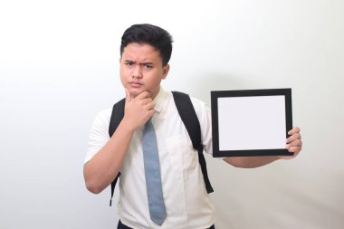 Endonezyalı lise öğrencisi beyaz gömlek giyiyor gri kravat takıyor ve kafasında kafa karışıklığı olan boş bir çerçeve gösteriyor. Tipografi ya da doldurulacak metin için boş boşluk