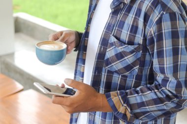 Asyalı adamın elinde bir fincan kahve ve cep telefonu tutarken yakın plan. Teknoloji ve sabah rutin konsepti